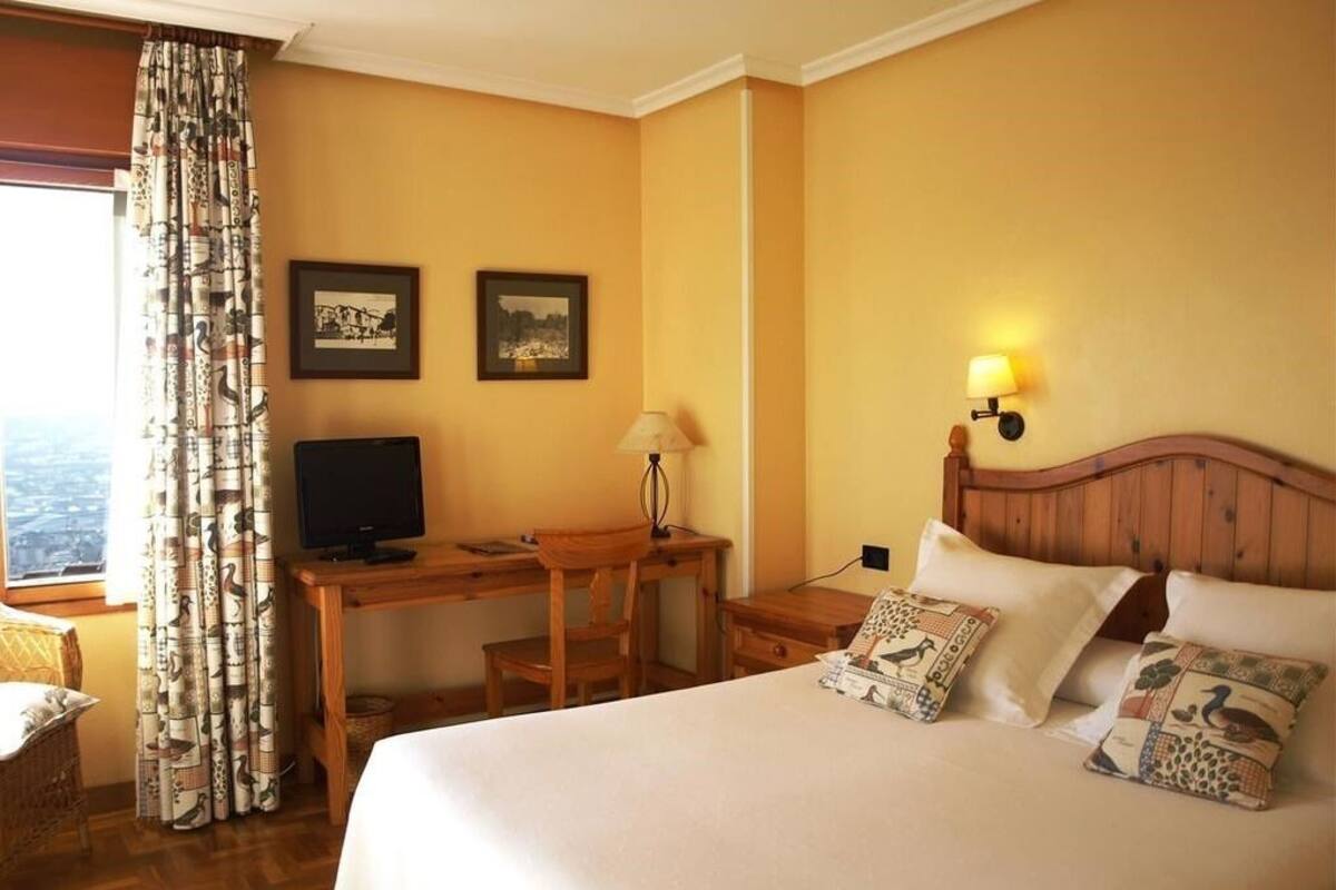 Quarto de hotel com uma cama, uma escrivaninha, uma televisão e duas molduras na parede.