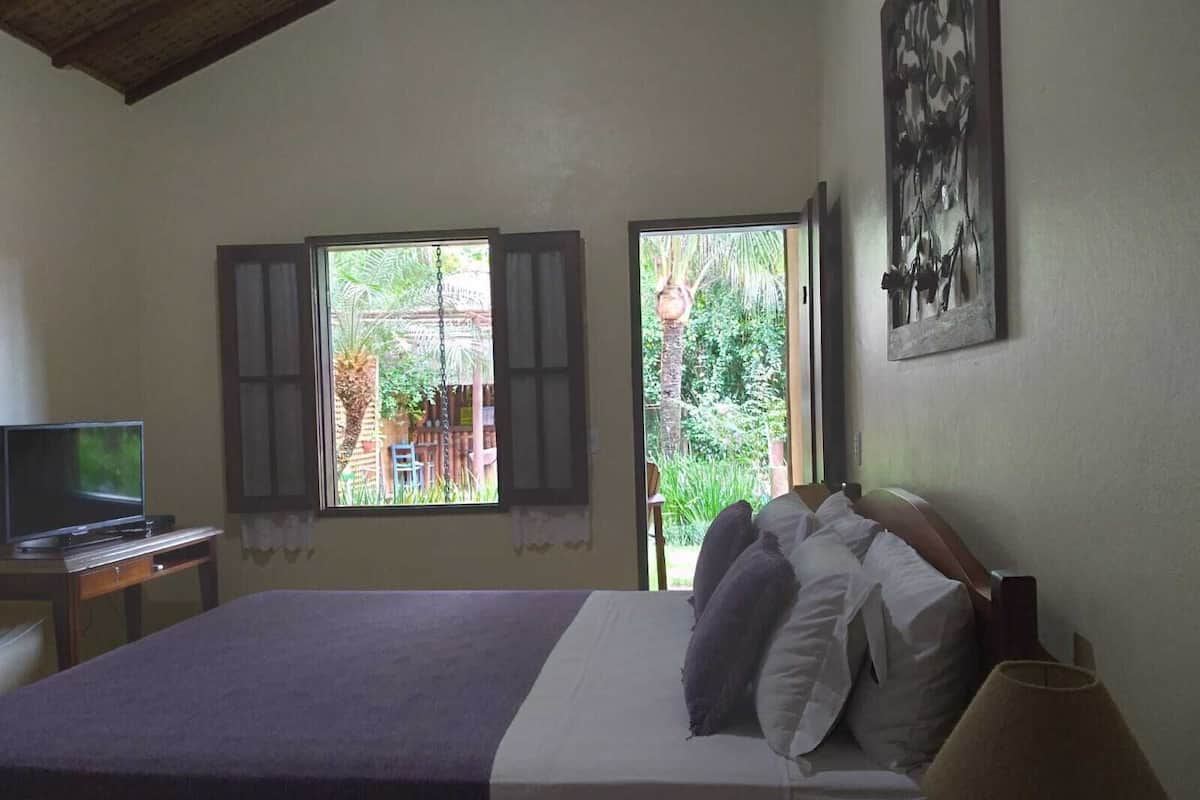 Ein Schlafzimmer mit einem Bett, einem Schreibtisch mit Computer, zwei Fenstern und einem Gemälde an der Wand.