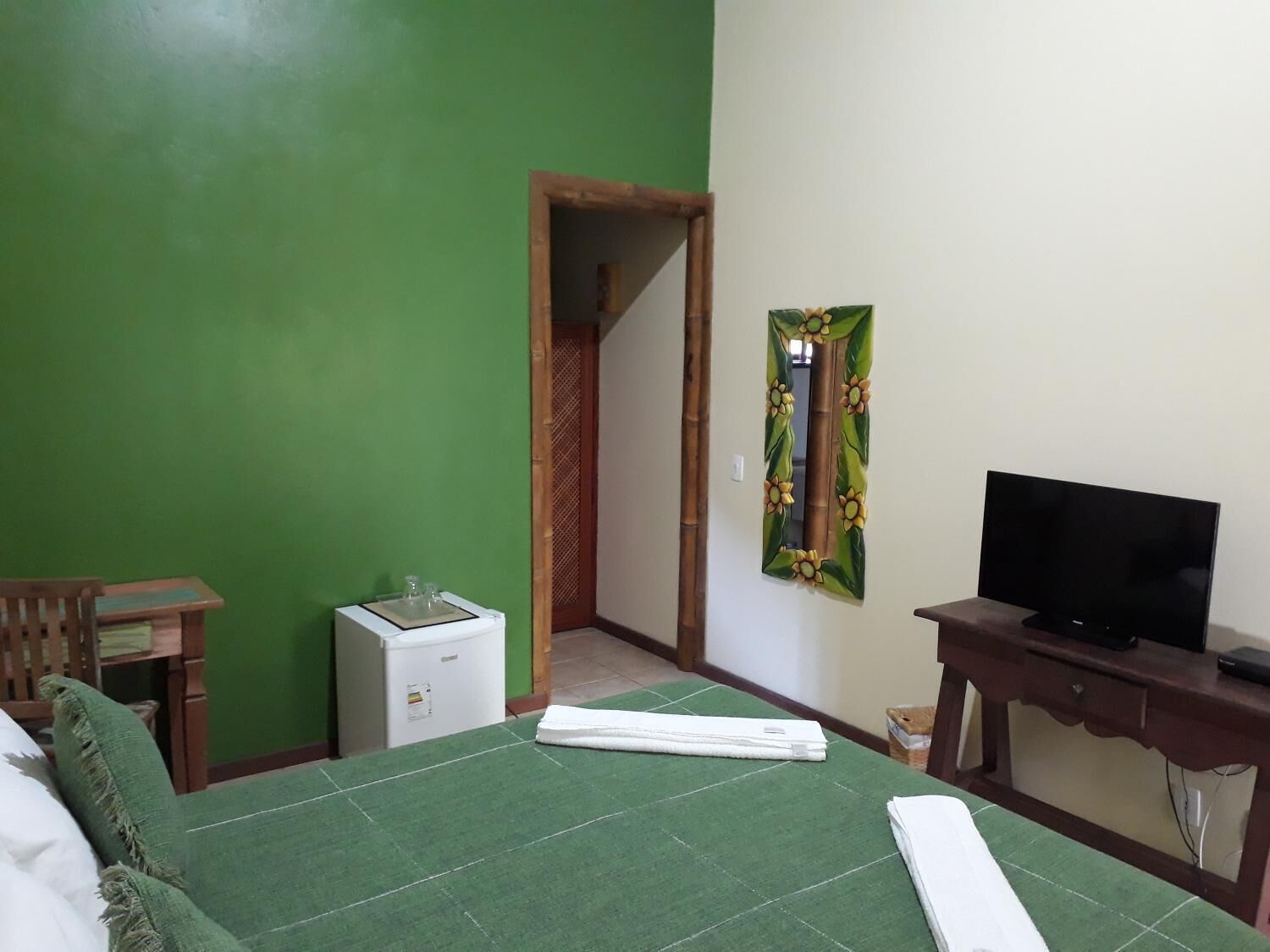 Une chambre d’hôtel avec un mur vert, un lit recouvert d’une couverture verte, un téléviseur posé sur un meuble en bois, un petit appareil électroménager blanc et un miroir orné de motifs floraux.