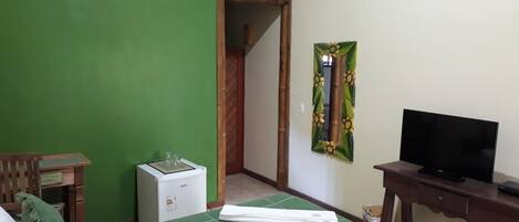 Une chambre d’hôtel avec un mur vert, un lit recouvert d’une couverture verte, un téléviseur posé sur un meuble en bois, un petit appareil électroménager blanc et un miroir orné de motifs floraux.