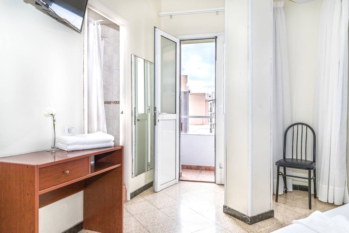 A hotel room with a wooden desk, a chair, a bed, and a view of the outside.