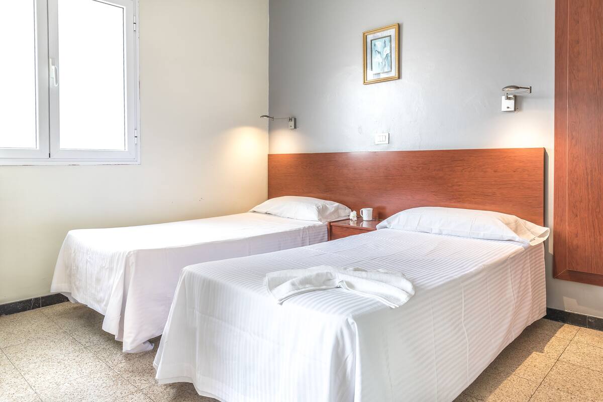A hotel room with two single beds, a wooden headboard, a small bedside table with a cup, and a framed picture on the wall.