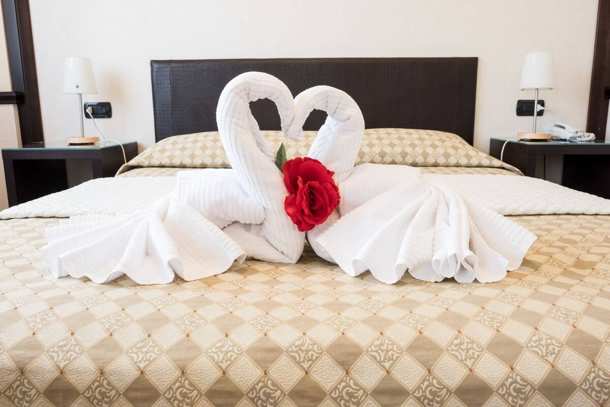 A neatly made bed with a diamond-patterned bedspread, two white towels arranged in swan shapes, and a red rose between them.