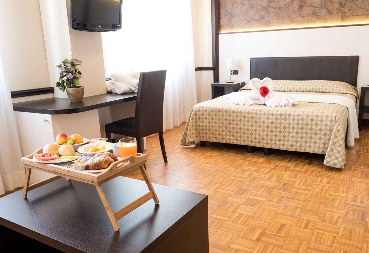 A hotel room with a bed, a desk with breakfast, a TV, and a potted plant.
