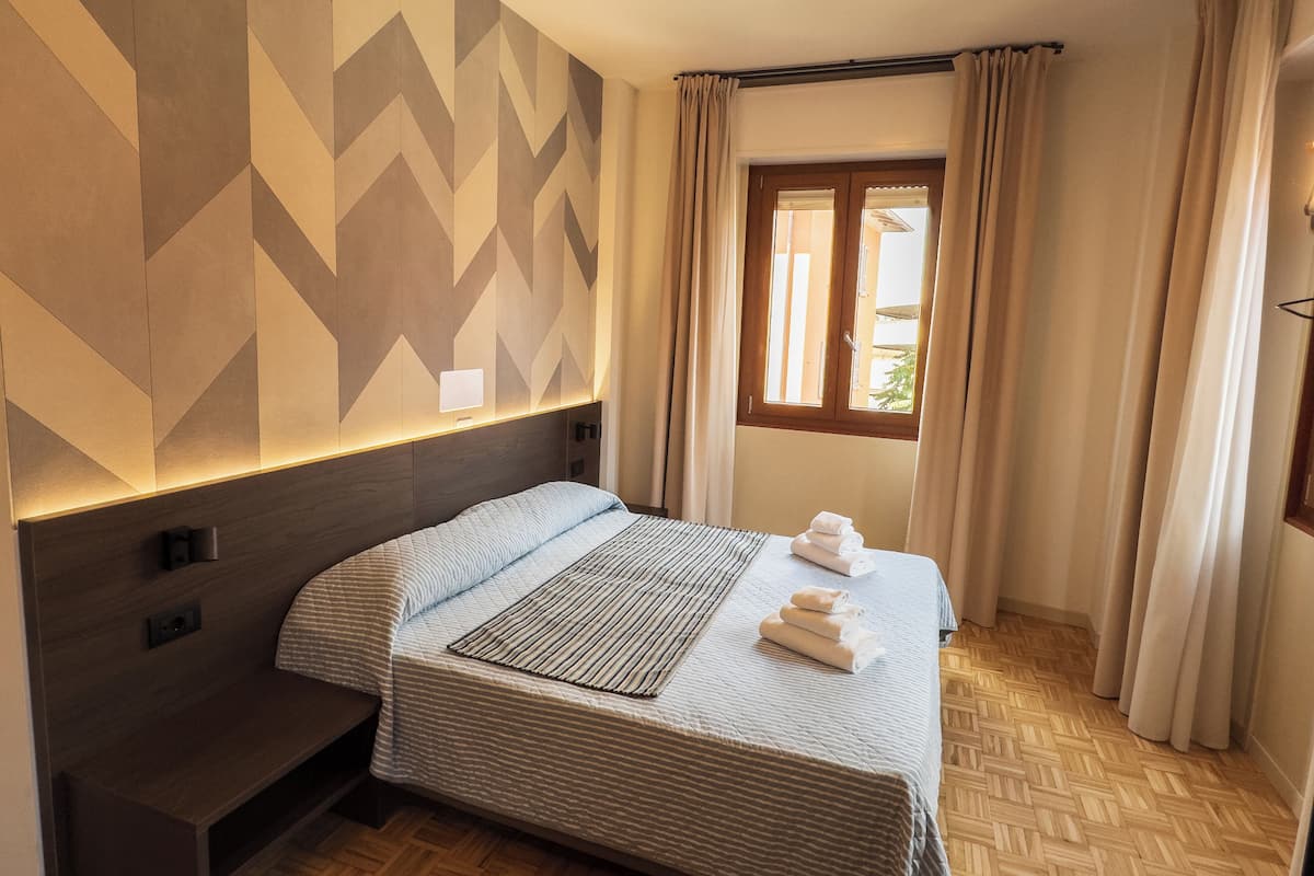 A hotel room with a bed, a bench, a wall with geometric patterns, a window with curtains, and a wooden floor.