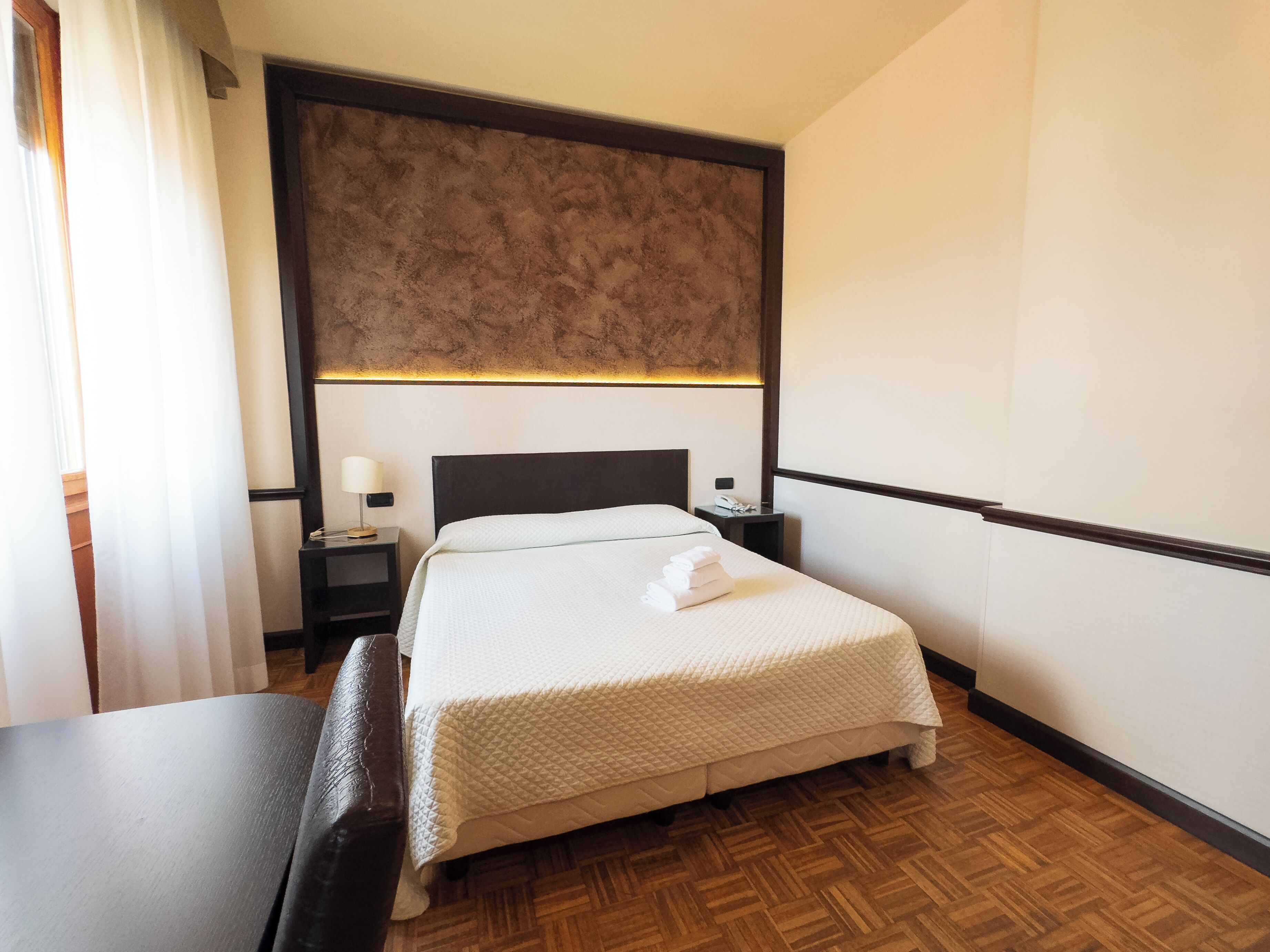 A hotel room with a bed, bedside table, lamp, and a wooden floor.