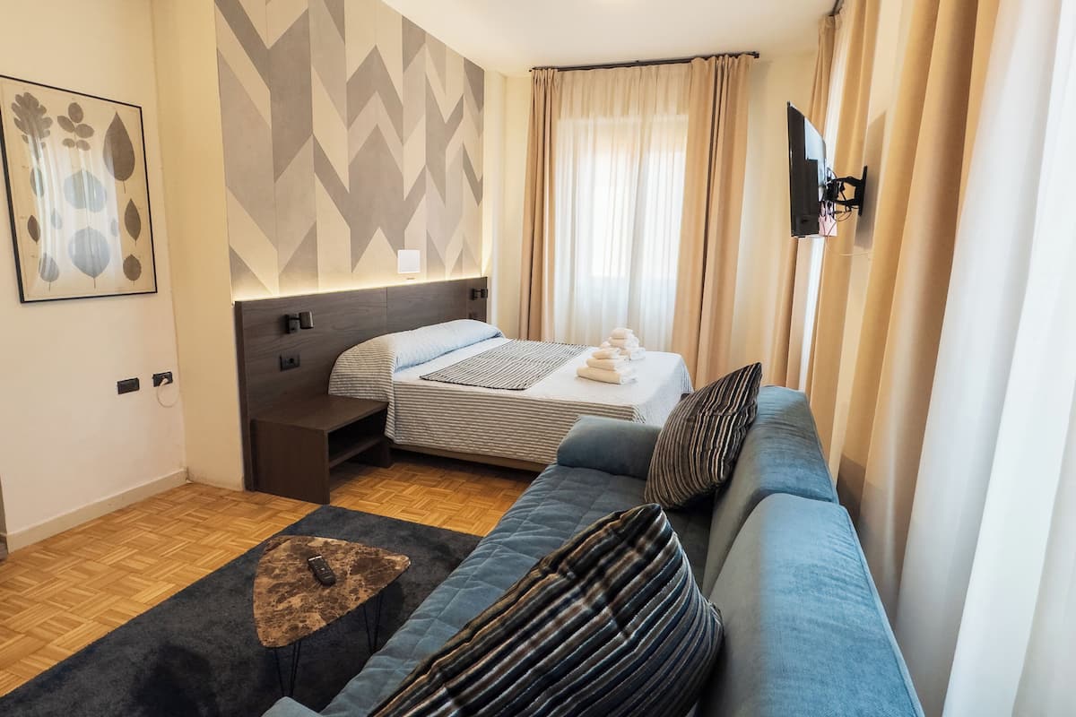 A modern hotel room with a sofa, a bed, a small table, and a wall-mounted TV.