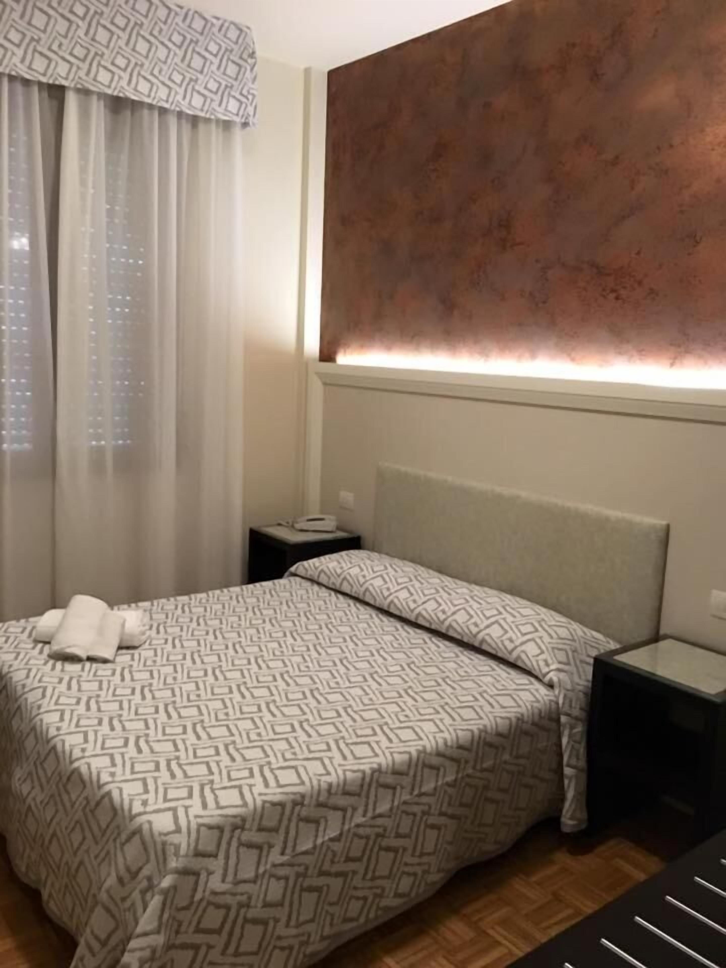 Una habitación de hotel con cama, mesita de noche, lámpara y ventana con cortinas.