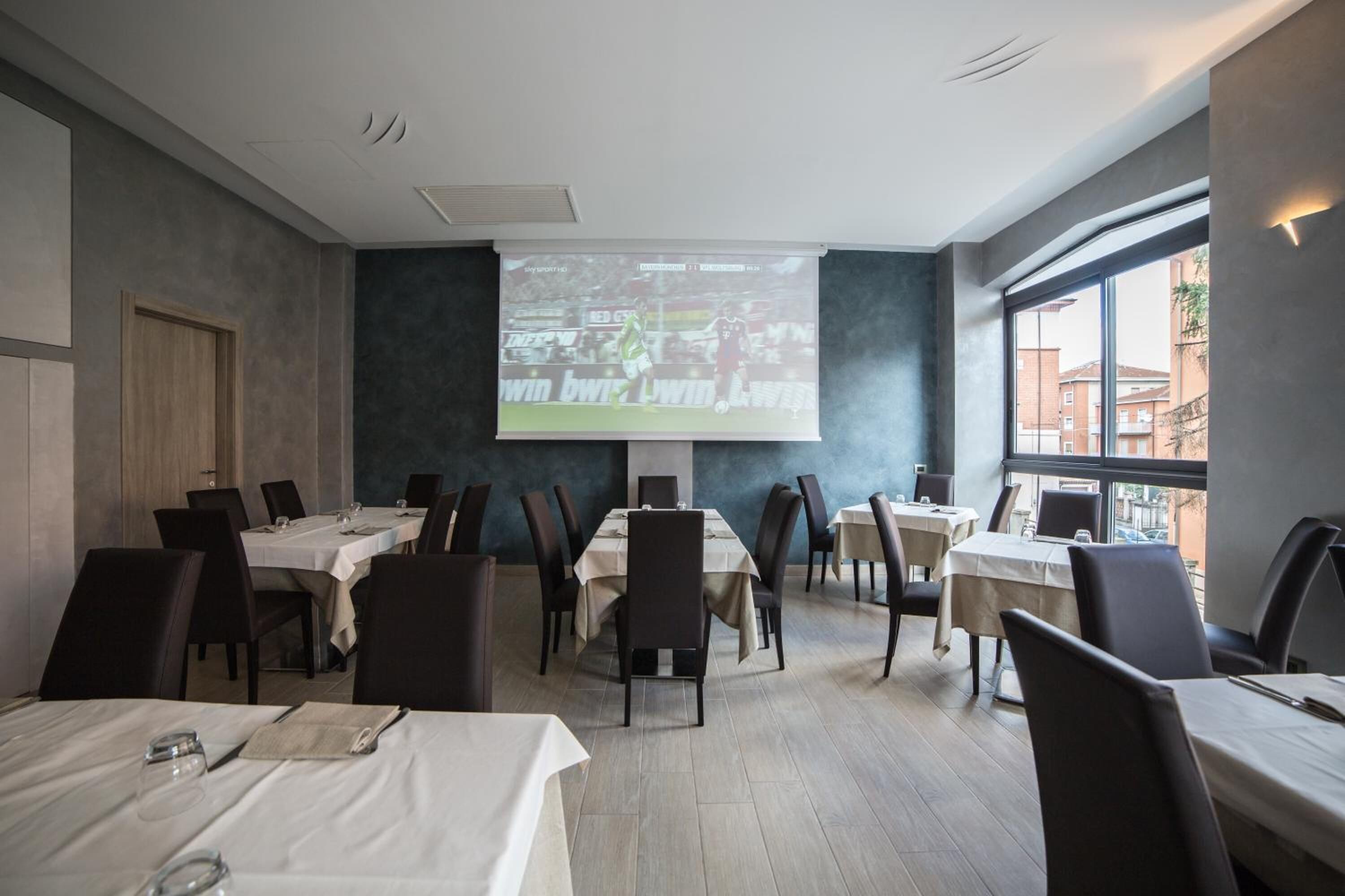 Ein Restaurant mit einer großen Projektionsleinwand, die ein Fußballspiel zeigt, Tische mit weißen Tischdecken und Blick auf Gebäude durch große Fenster.