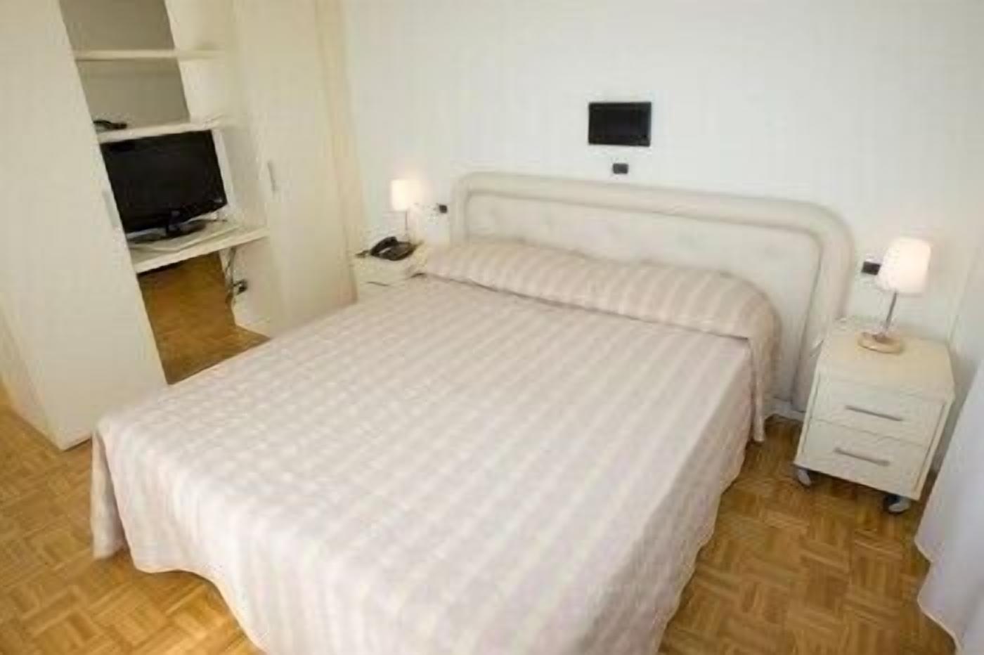 Ein Zimmer mit einem Einzelbett, Holzboden, einem Bett mit gestreifter Bettdecke, einem Nachttisch mit Lampe sowie einem Fernseher auf einem Regal.