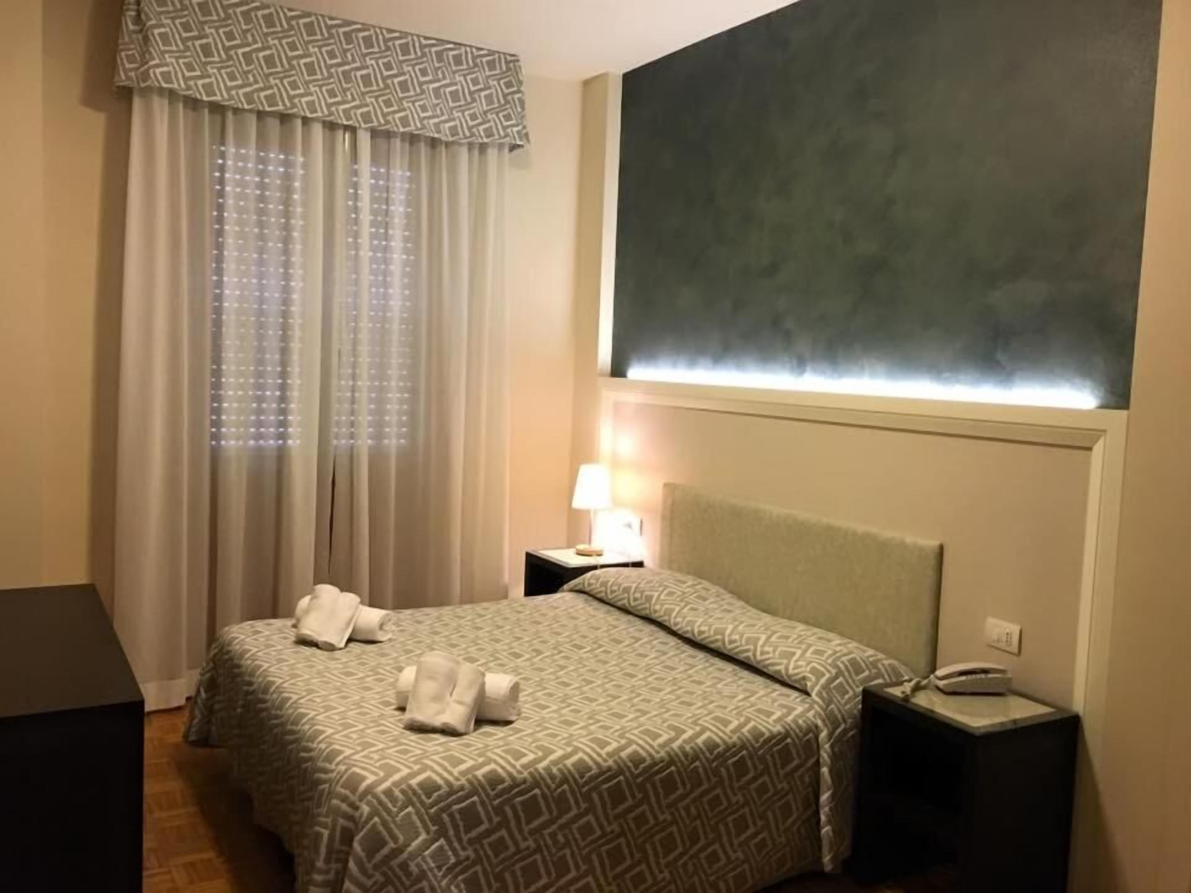 A hotel room with a bed, a nightstand, a lamp, and a large window with curtains.