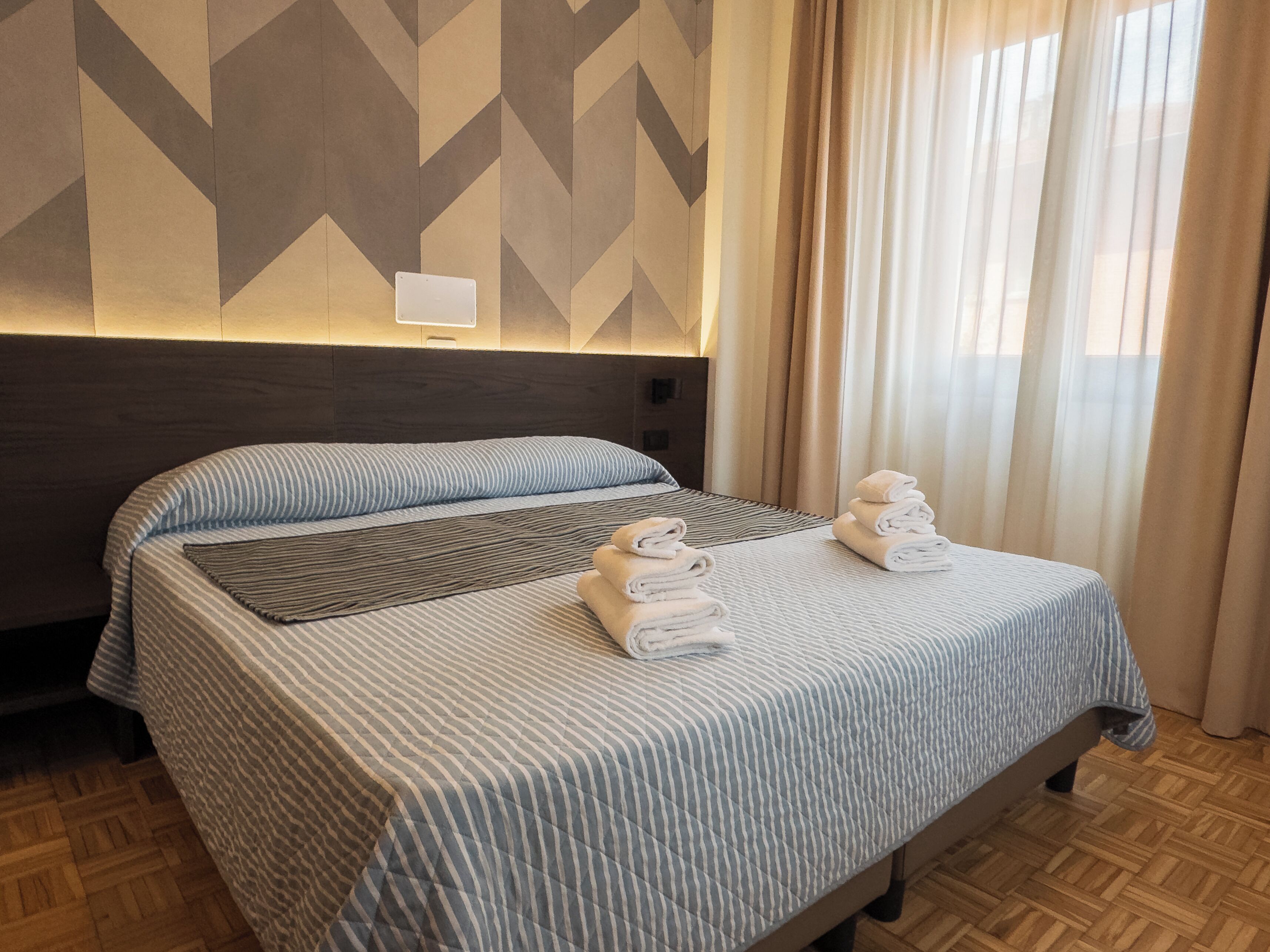 Habitación de hotel con cama, cabecero, pared con diseño geométrico y ventana con cortinas.