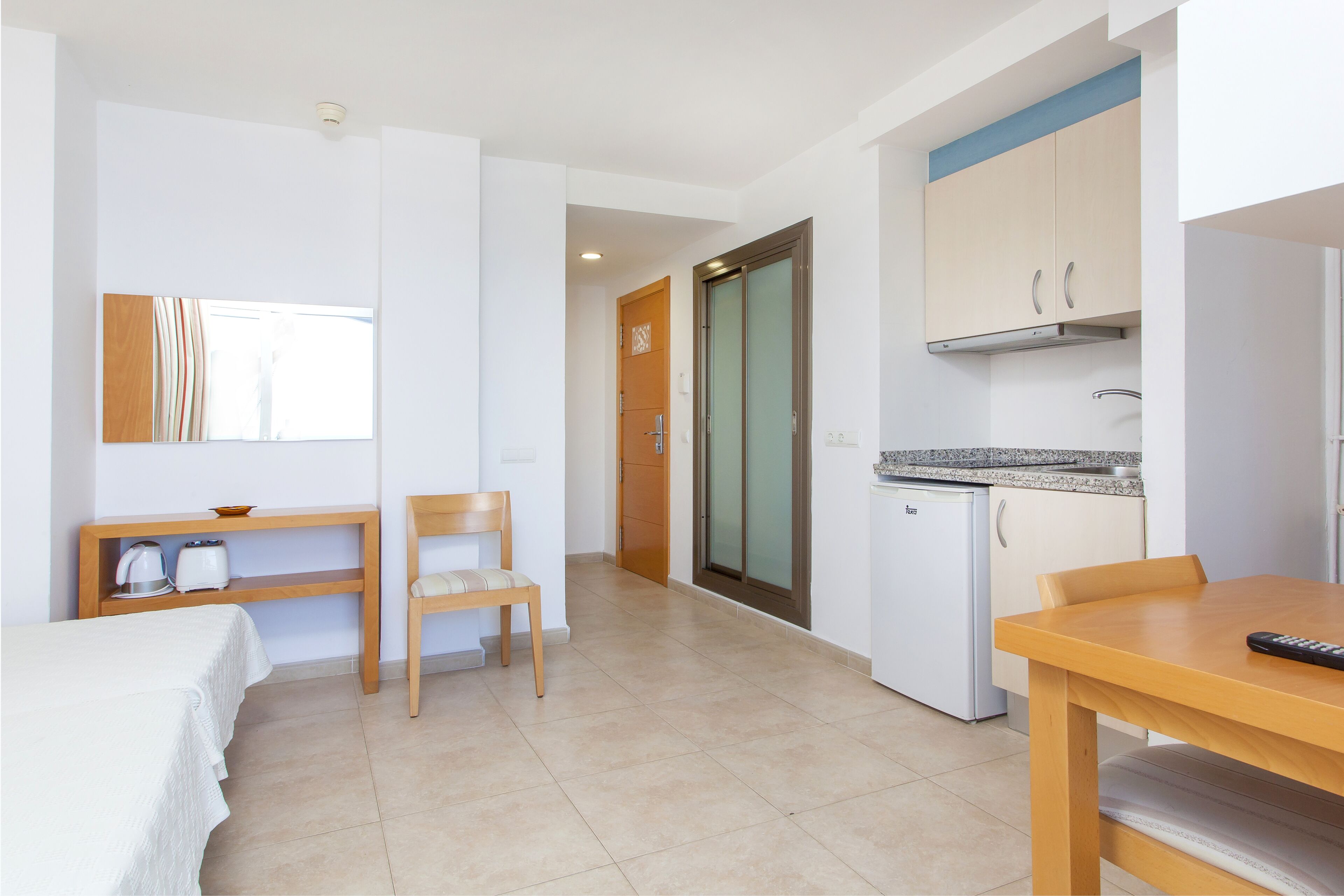 A hotel room with a bed, a wooden chair, a small table, a kitchenette, and a door leading to another room.