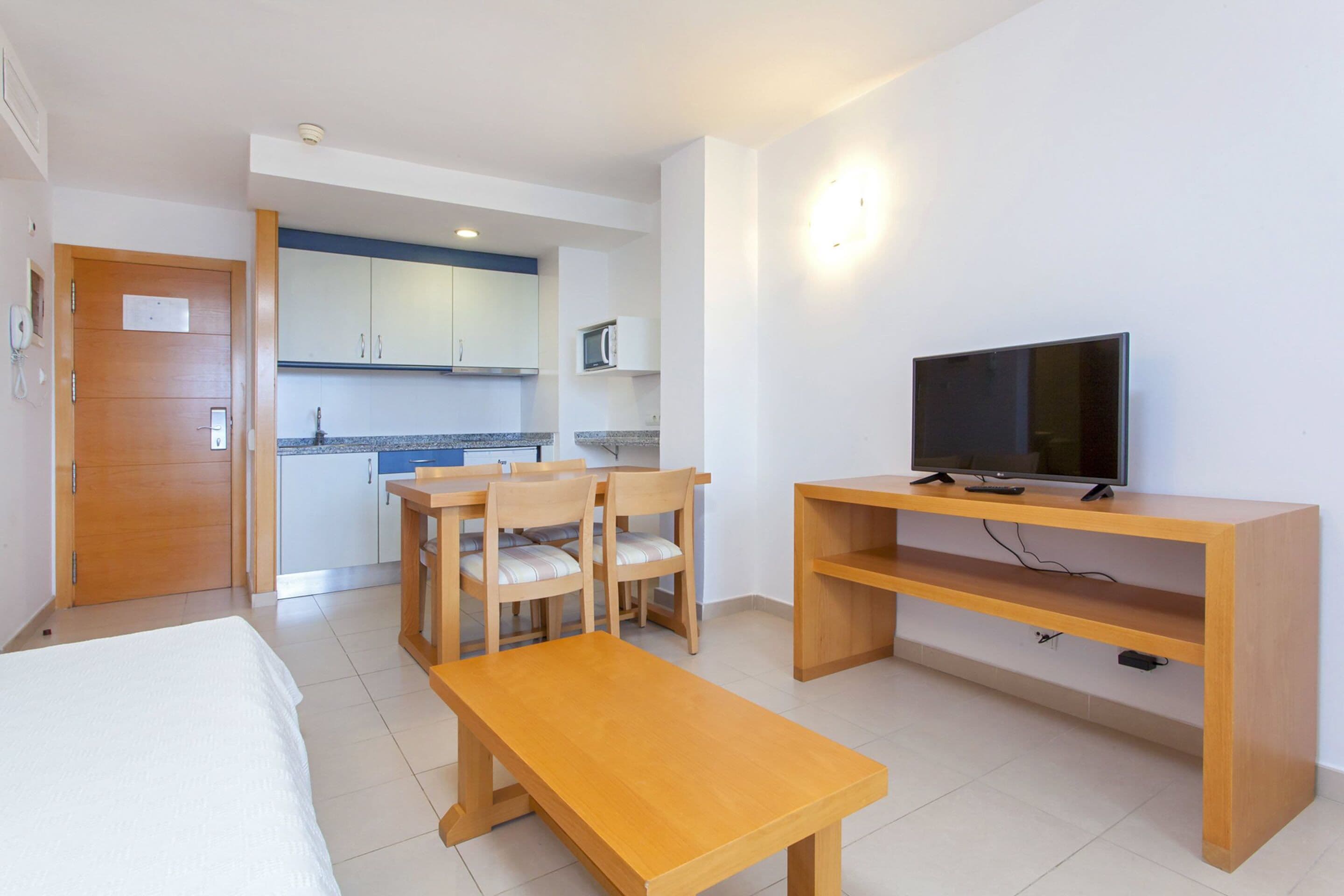 A hotel room with a bed, a wooden table, a TV on a stand, and a kitchenette in the background.