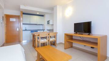 Habitación de hotel con cama, una mesa de madera, televisor sobre un mueble bajo y una kitchenette al fondo.
