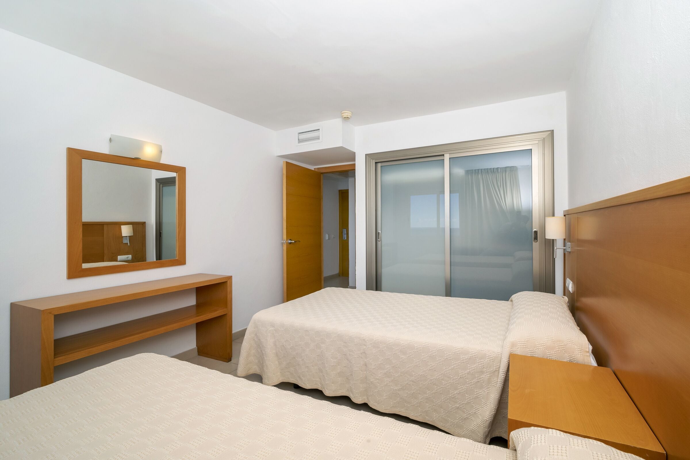 Habitación de hotel con dos camas, una mesita consola de madera, un espejo, una puerta corrediza de vidrio y un escritorio pequeño.
