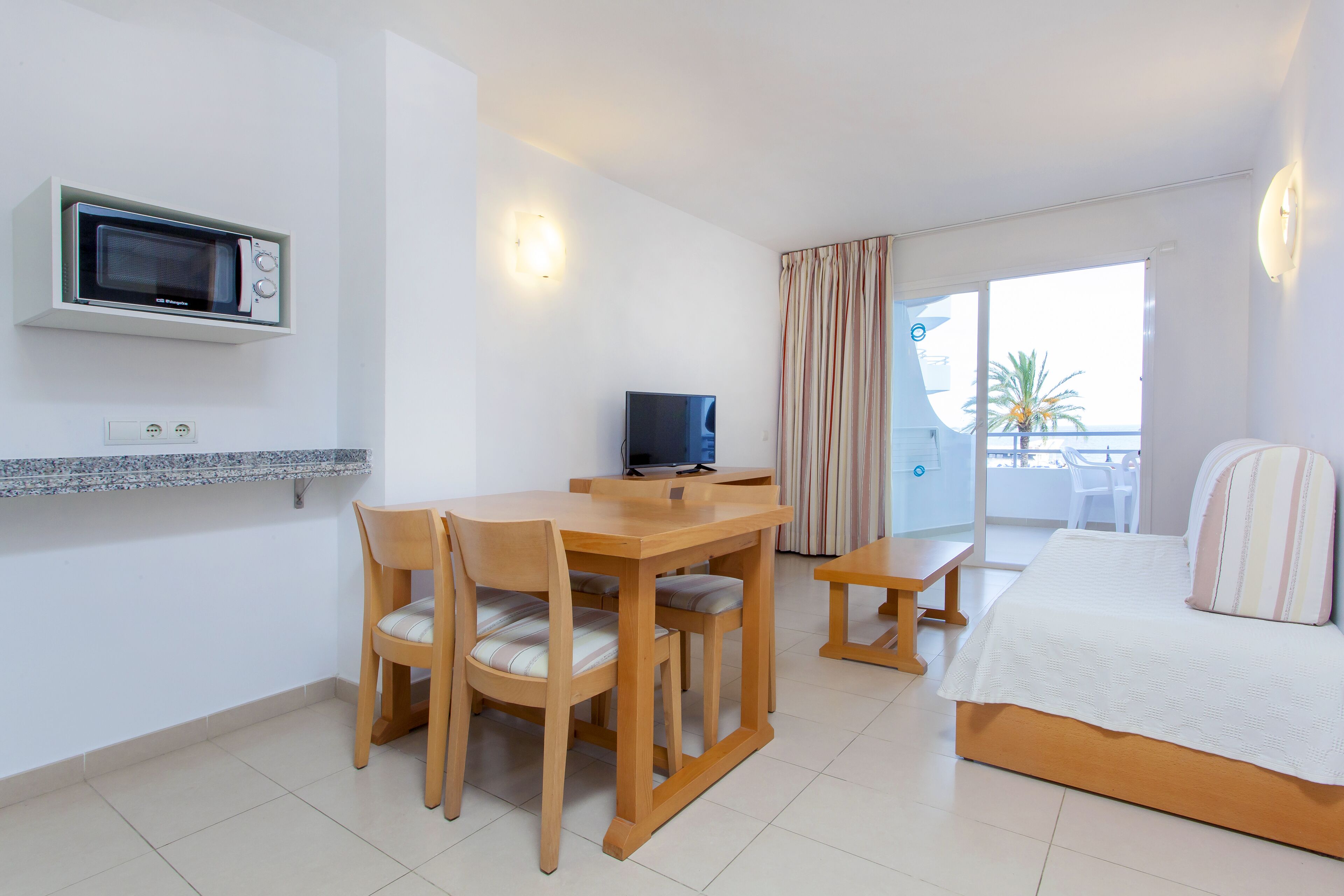 A hotel room with a wooden dining table, chairs, a TV, a bed, and a balcony with curtains.