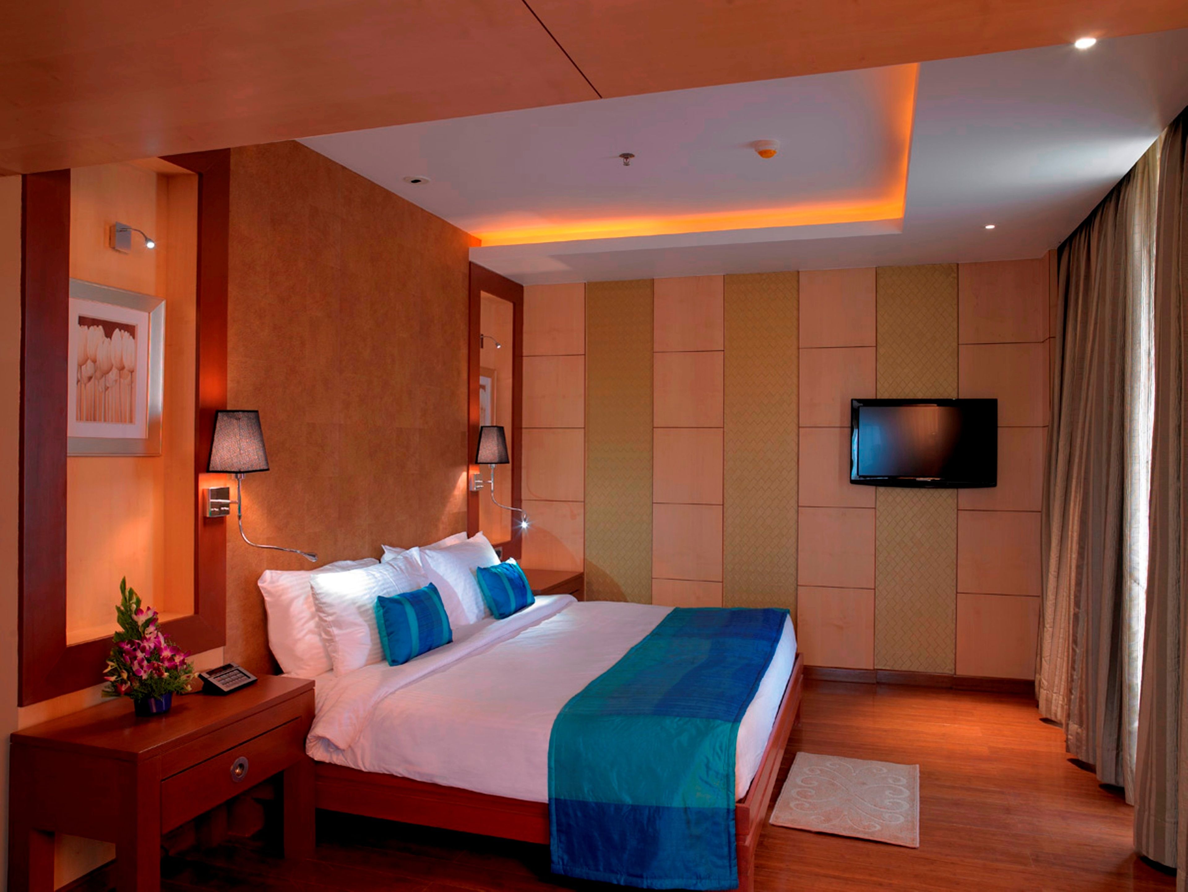 A hotel room with a large bed, a flat-screen TV mounted on the wall, a nightstand with a lamp, and a small table with a flower vase.