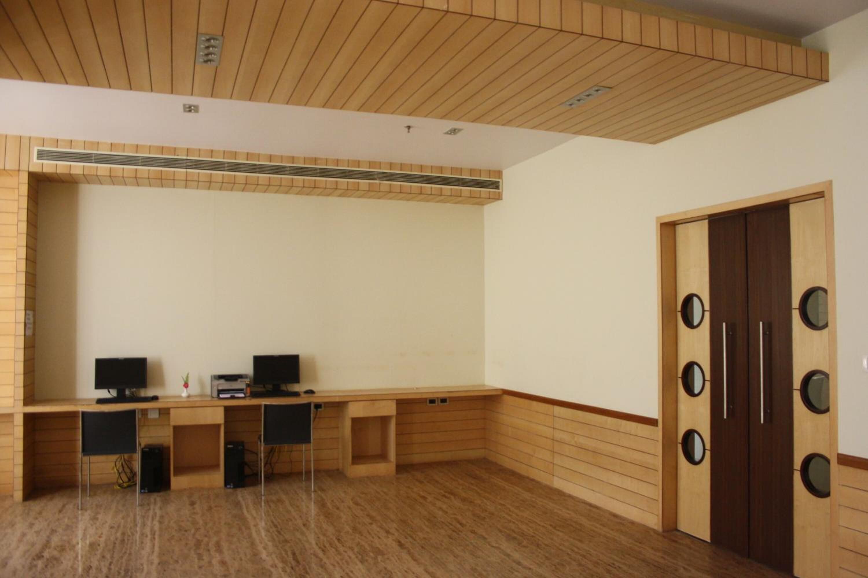 A room with a long wooden desk, two monitors, and a printer. There are two chairs, a cabinet, and a door with circular mirrors.