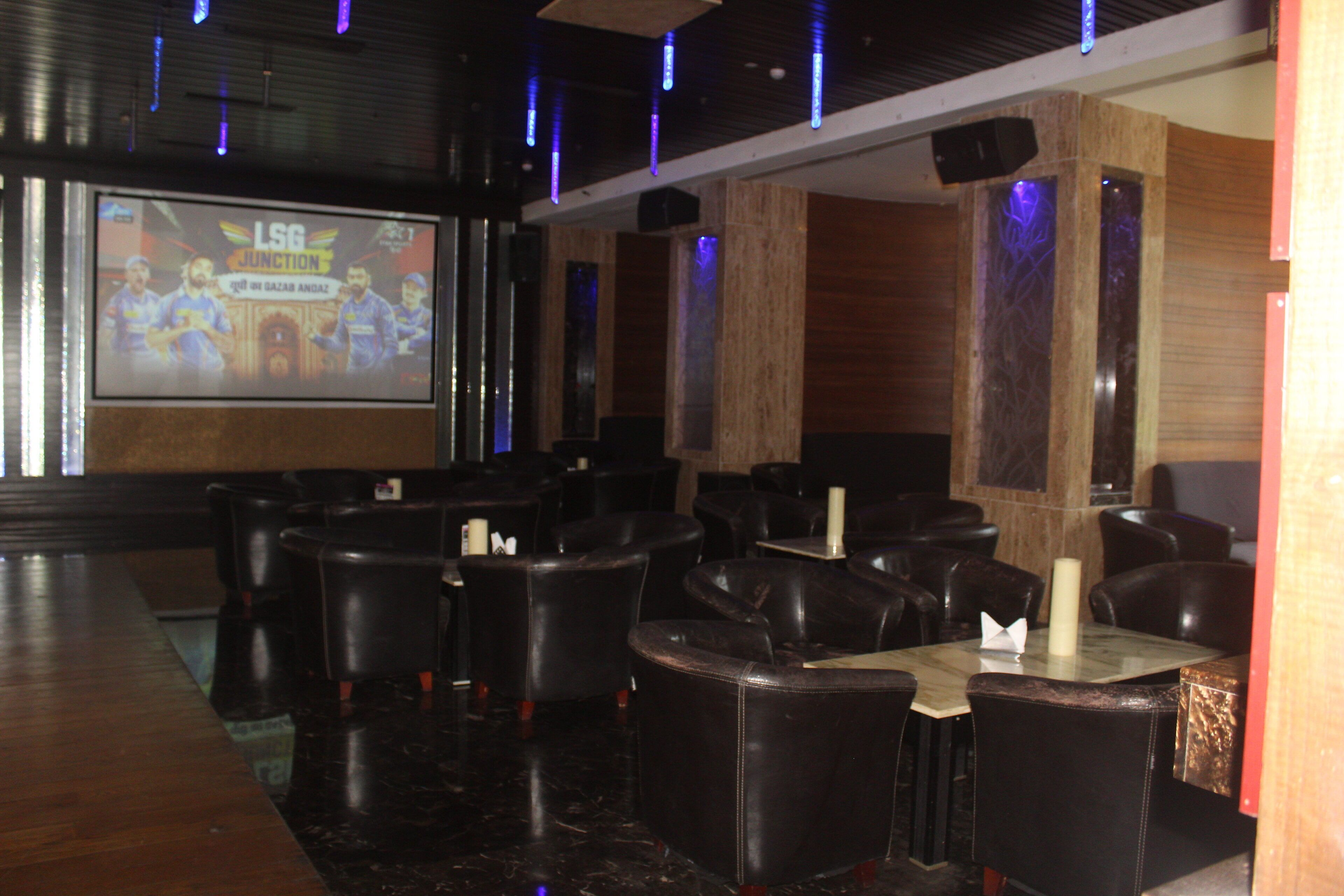 A restaurant with black leather chairs and tables, a large screen displaying a sports event, and a wooden accent wall.