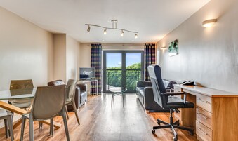 A modern hotel room with a flat-screen TV, a desk, a chair, a couch, and a balcony with a view of greenery.