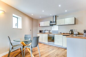 A modern kitchen with white cabinets, a microwave, oven, and a coffee maker. There is a dining table with chairs and a window providing natural light.