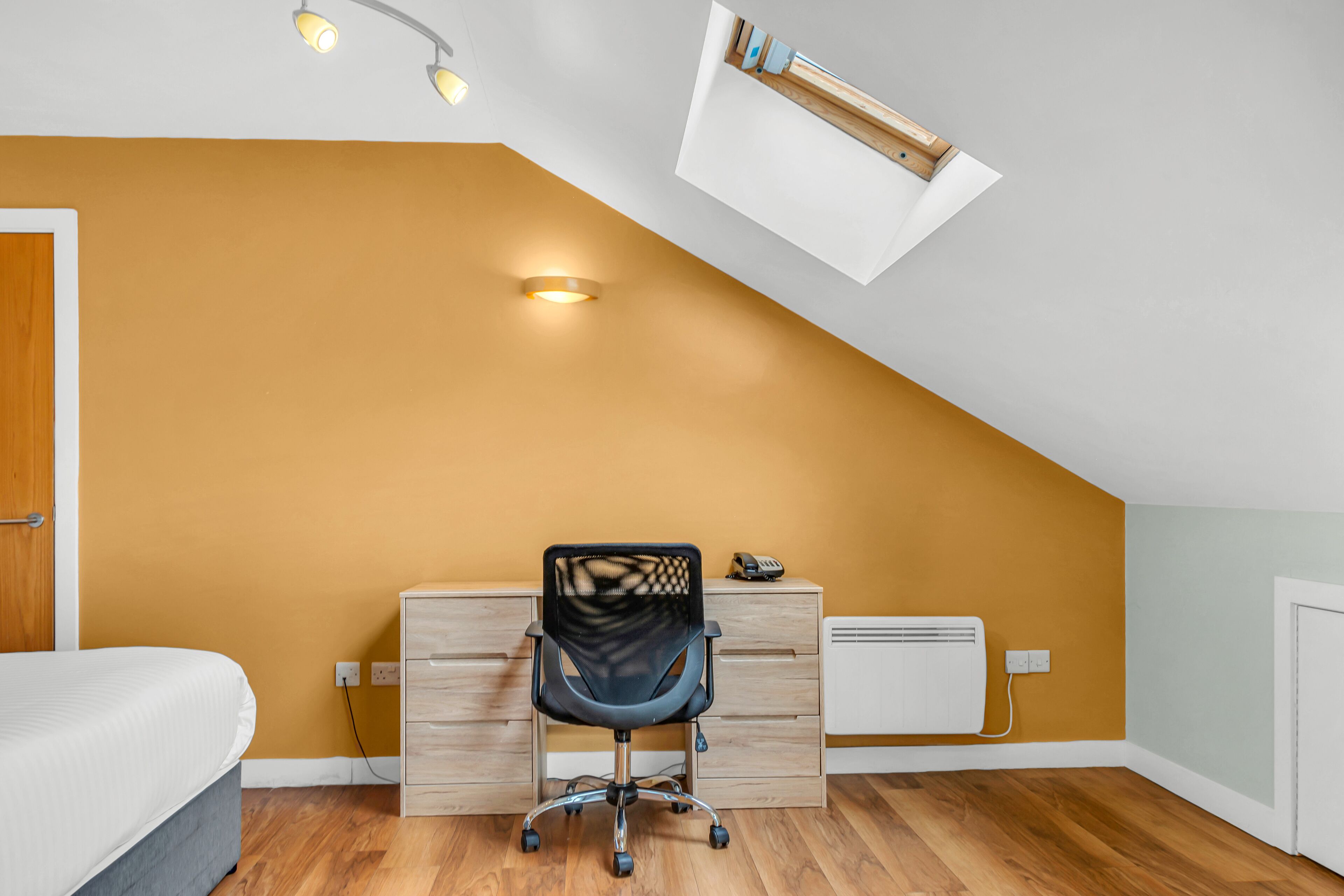 Een slaapkamer met een schuine kap, een bureau met lades, een stoel en een bed.