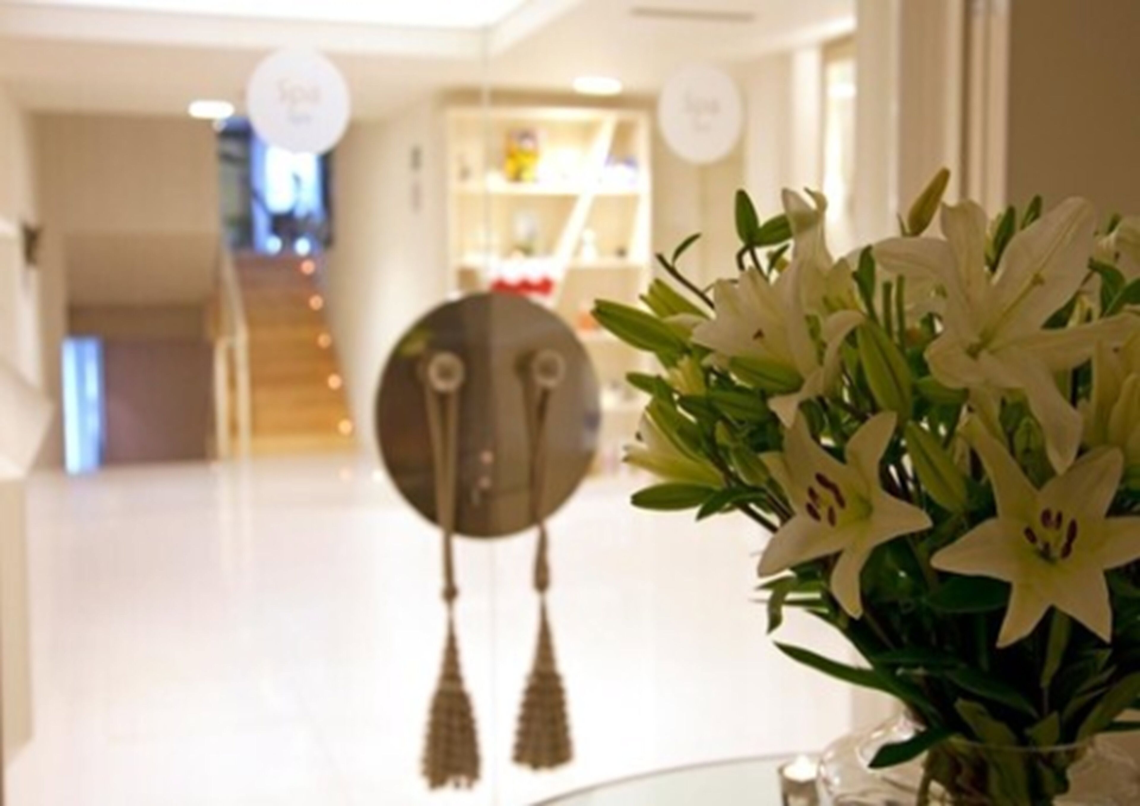 Un hall de hotel con mostrador de recepción, un espejo redondo con borlas y un jarrón de lirios blancos.