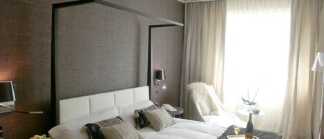 A hotel room with a large bed, a flat-screen TV, a bedside table with a lamp, and a view of the outdoors through the window.