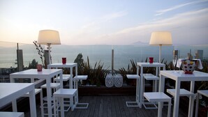Een terras met witte tafels en stoelen, uitzicht op zee en decoratieve planten.