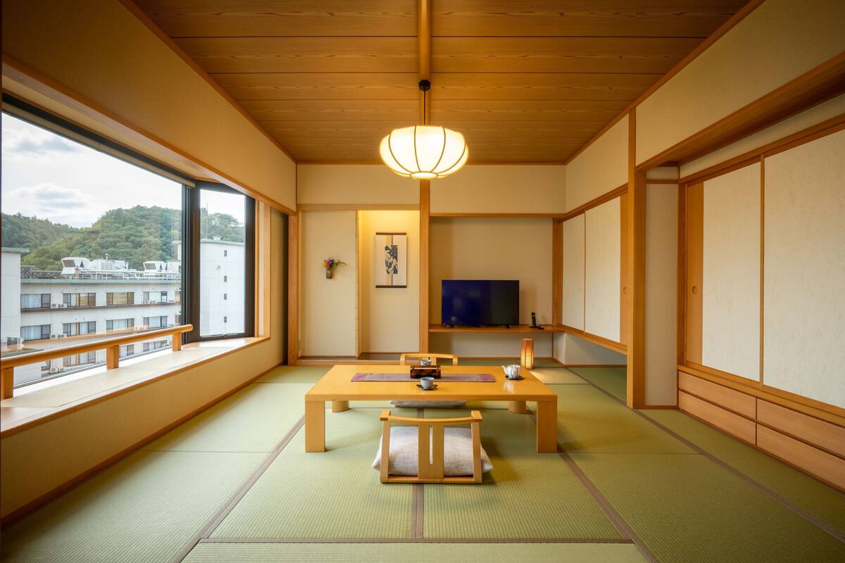 Ett traditionellt japanskt rum med tatamimattor på golvet, ett lågt träbord, en tv och ett stort fönster med utsikt över ett bostadsområde.