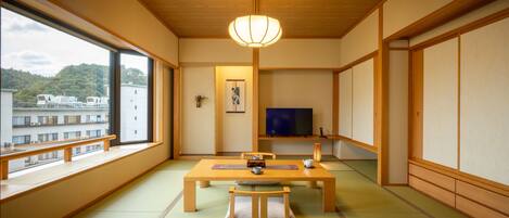 A traditional Japanese room with tatami flooring, a low wooden table, a television, and a large window overlooking a residential area.