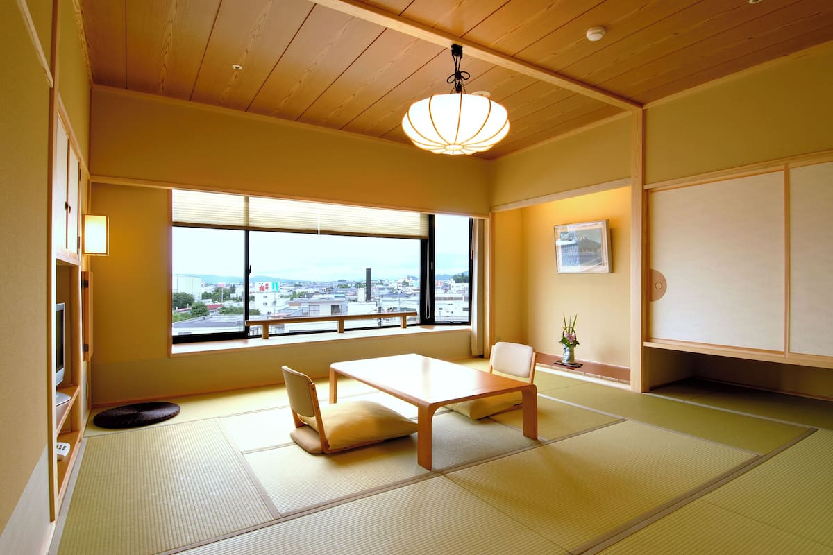 Ett traditionellt japanskt rum med tatamimattor på golvet, ett lågt bord, stolar, en hängande lampa, ett stort fönster med utsikt över staden och en inramad tavla på väggen.
