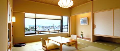 Una habitación tradicional japonesa con suelo de tatami, una mesa baja, sillas, una lámpara colgante, un amplio ventanal con vista a la ciudad y un cuadro en la pared.
