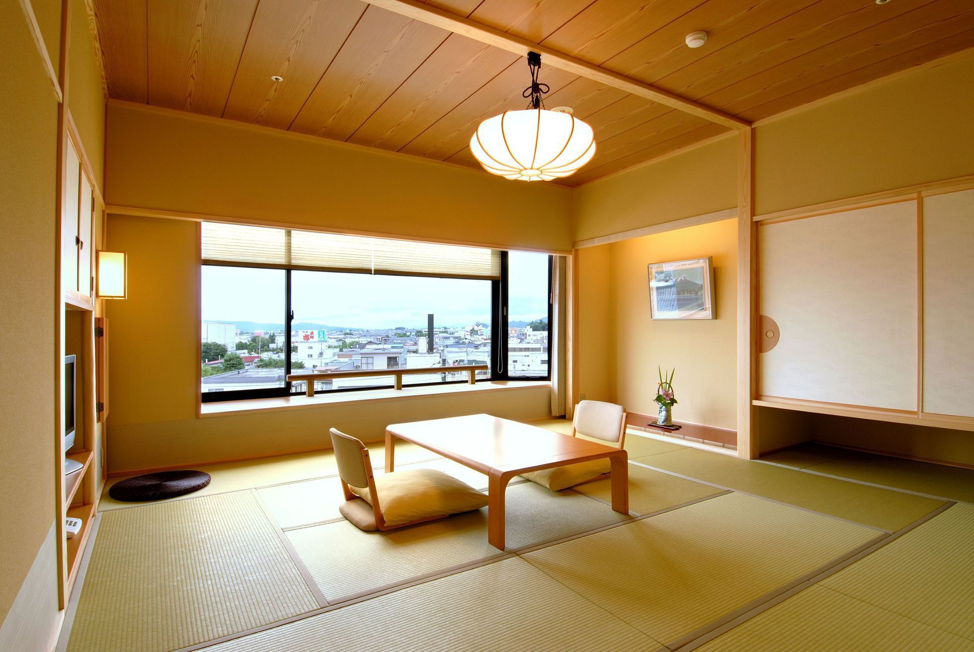 A traditional Japanese room with tatami flooring, a low table, a chair, a hanging light fixture, a window with a city view, and a sliding door.