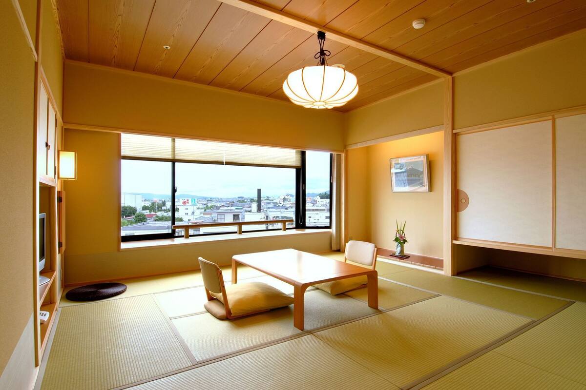 A traditional Japanese room with tatami flooring, a low table, a chair, a hanging light fixture, a window with a city view, and a sliding door.
