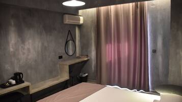 A modern hotel room with a bed, desk, and chair. There is an air conditioning unit on the wall and a curtain covering the window.