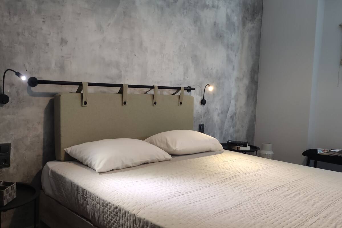 A bed with a headboard, two pillows, a bedside table with a lamp, and a wall-mounted light fixture.