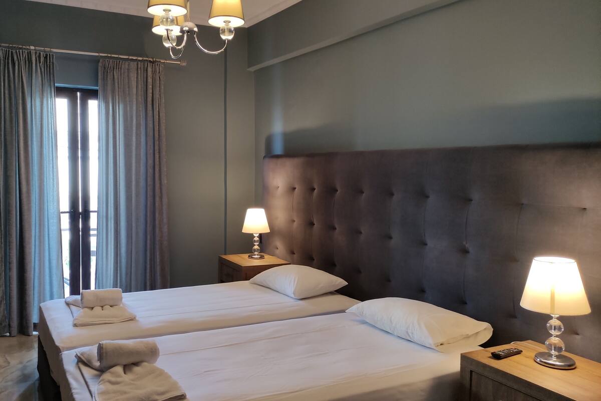 A hotel room with two beds, a large tufted headboard, a window with curtains, and a chandelier.