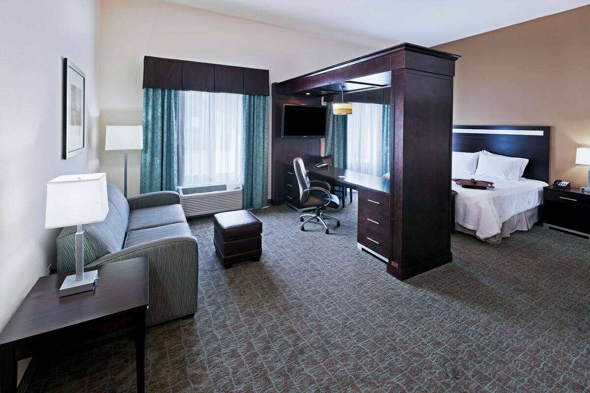 A hotel room with a bed, a desk with a chair, a sofa, a TV, and a lamp.