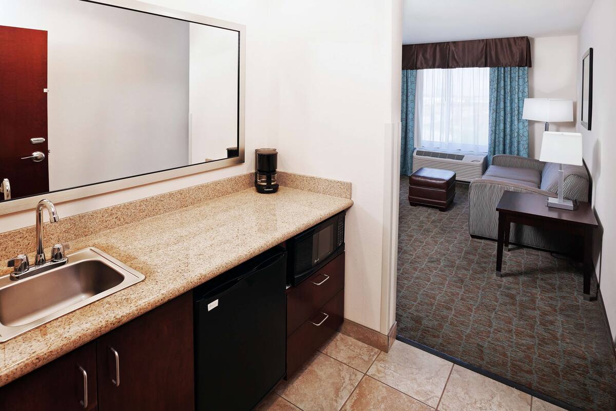 A hotel room with a kitchen area including a sink, microwave, and coffee maker. There is a seating area with a sofa and a table.