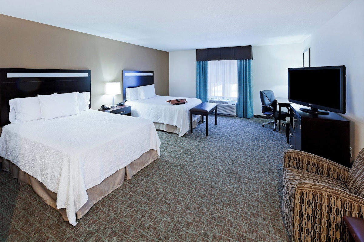 A hotel room with two beds, a desk, a chair, a television, and a large window with curtains.