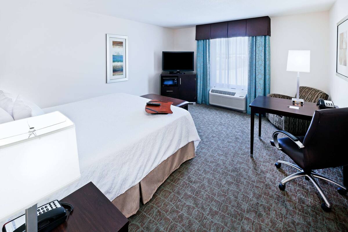 A hotel room with a bed, a desk, a chair, a television, a phone, and an air conditioning unit.