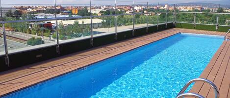 Una piscina con cubierta de madera y barandales de metal, rodeada por un paisaje urbano y vegetación.
