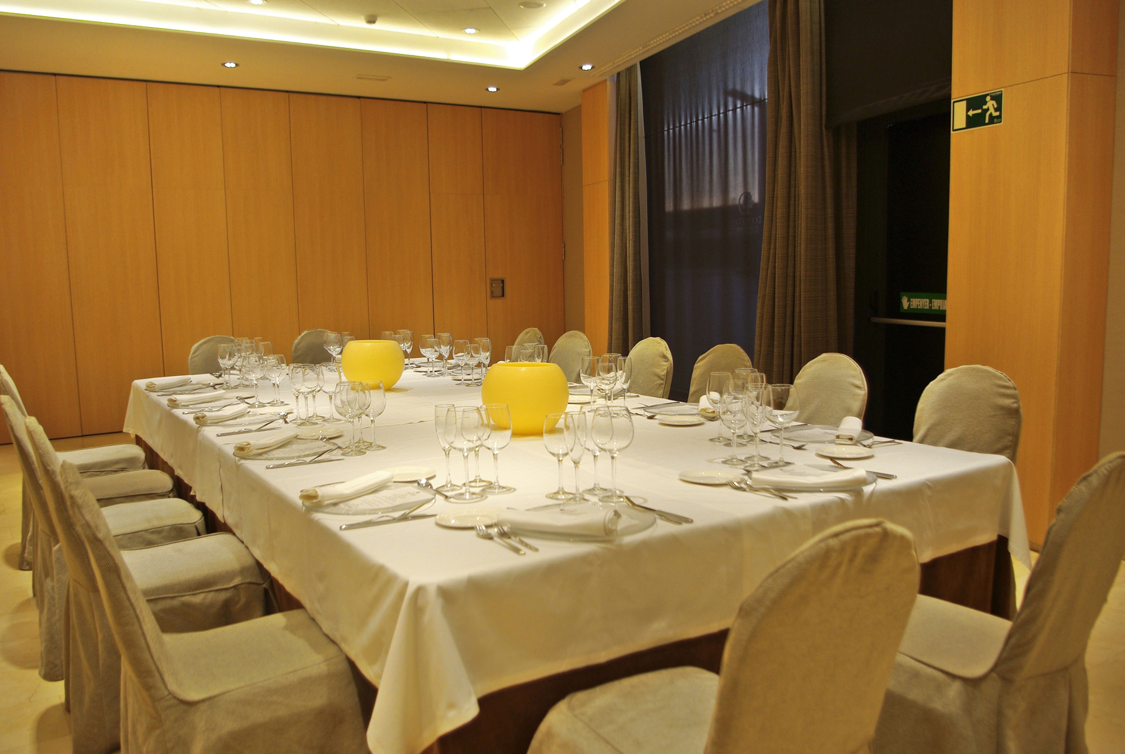 Un salón de conferencias con una larga mesa dispuesta para una reunión, equipado con sillas y un gran jarrón amarillo.