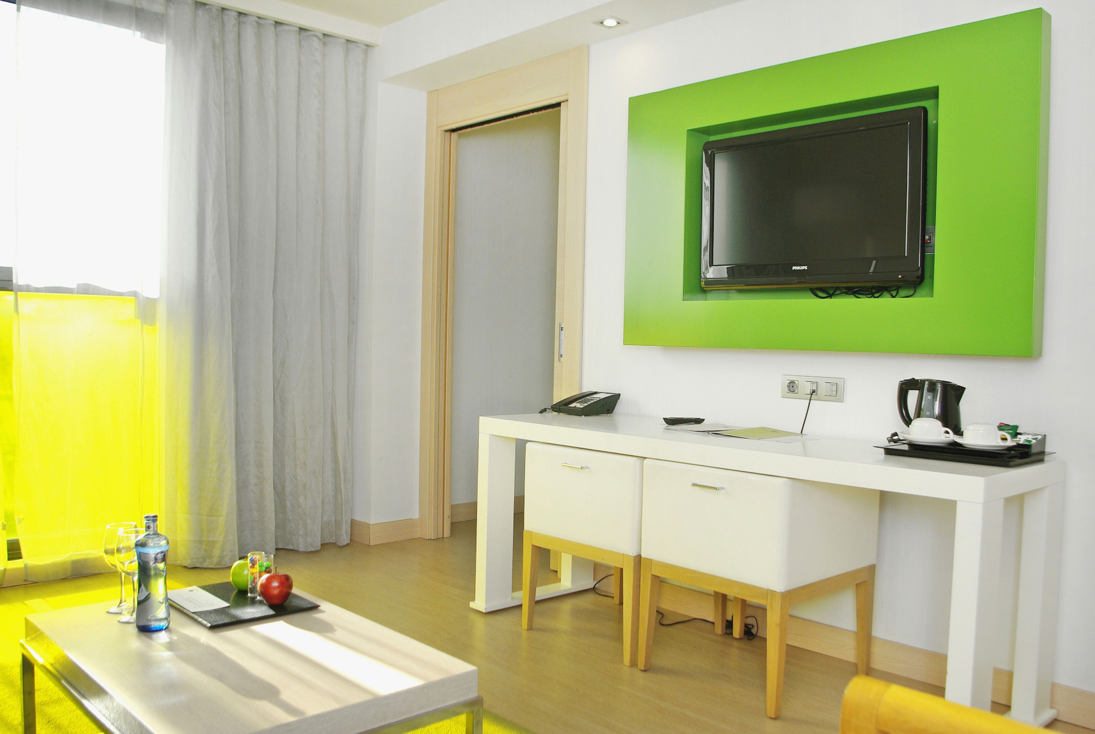 A modern hotel room with a white desk, a green-framed TV, a coffee maker, and a bottle of water on the table.
