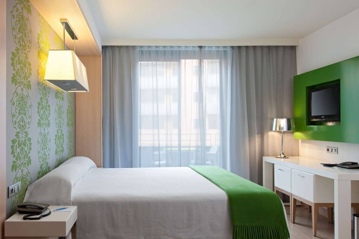 Habitación de hotel con cama, mesita de noche, escritorio, televisor y ventana con cortinas.