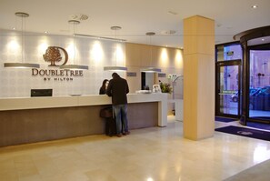 A hotel reception desk with a DoubleTree logo, a man standing at the counter, and a glass door entrance. - DoubleTree by Hilton Hotel Girona (Girona)