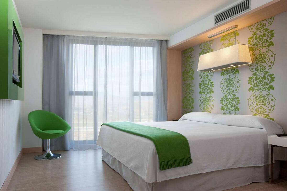 Habitación de hotel con cama, una silla, un escritorio, ventana con cortinas y pared con estampado floral.