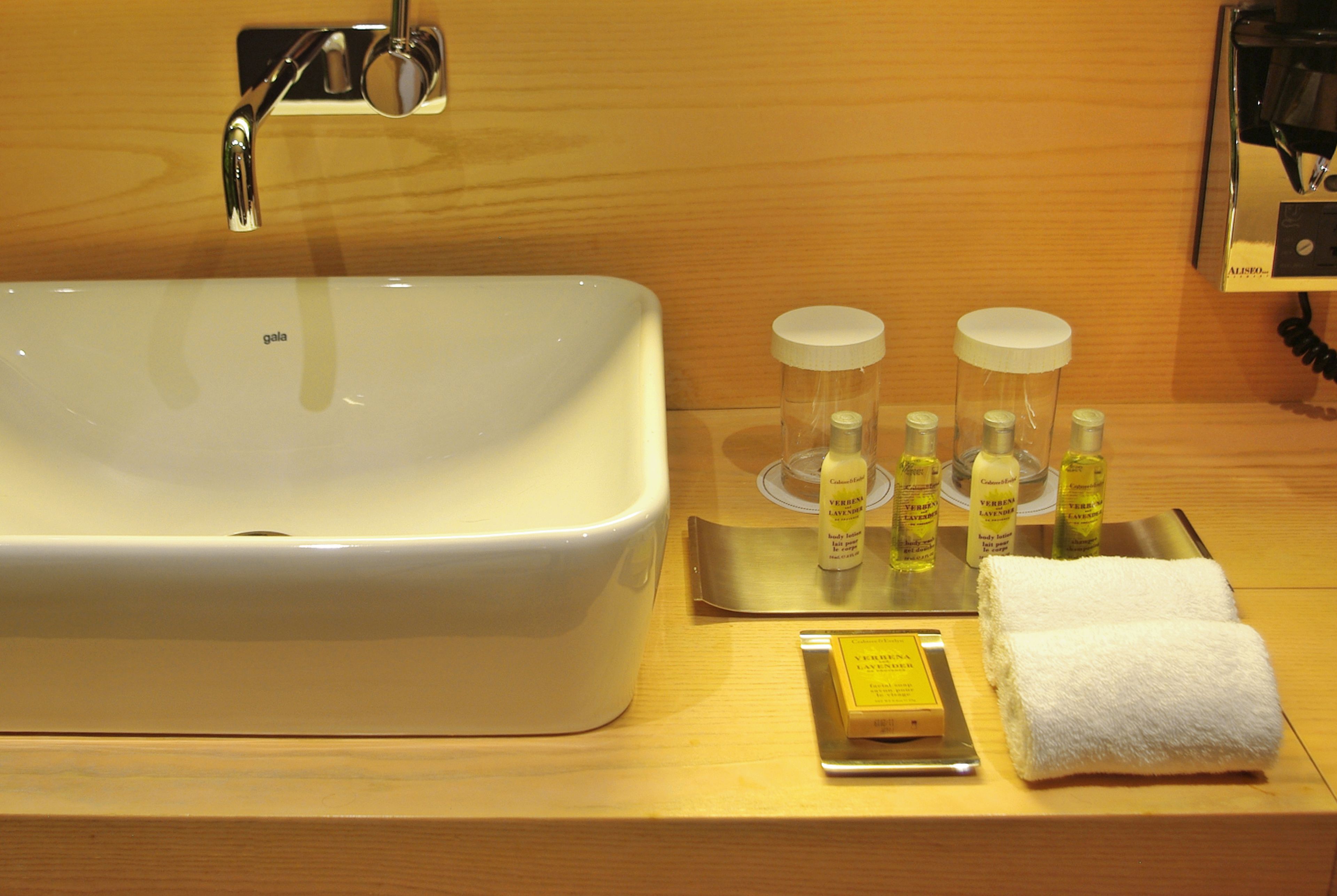 Un baño de hotel con un lavamanos blanco grande, un espejo y artículos de aseo, incluyendo botellas de loción y una pastilla de jabón.